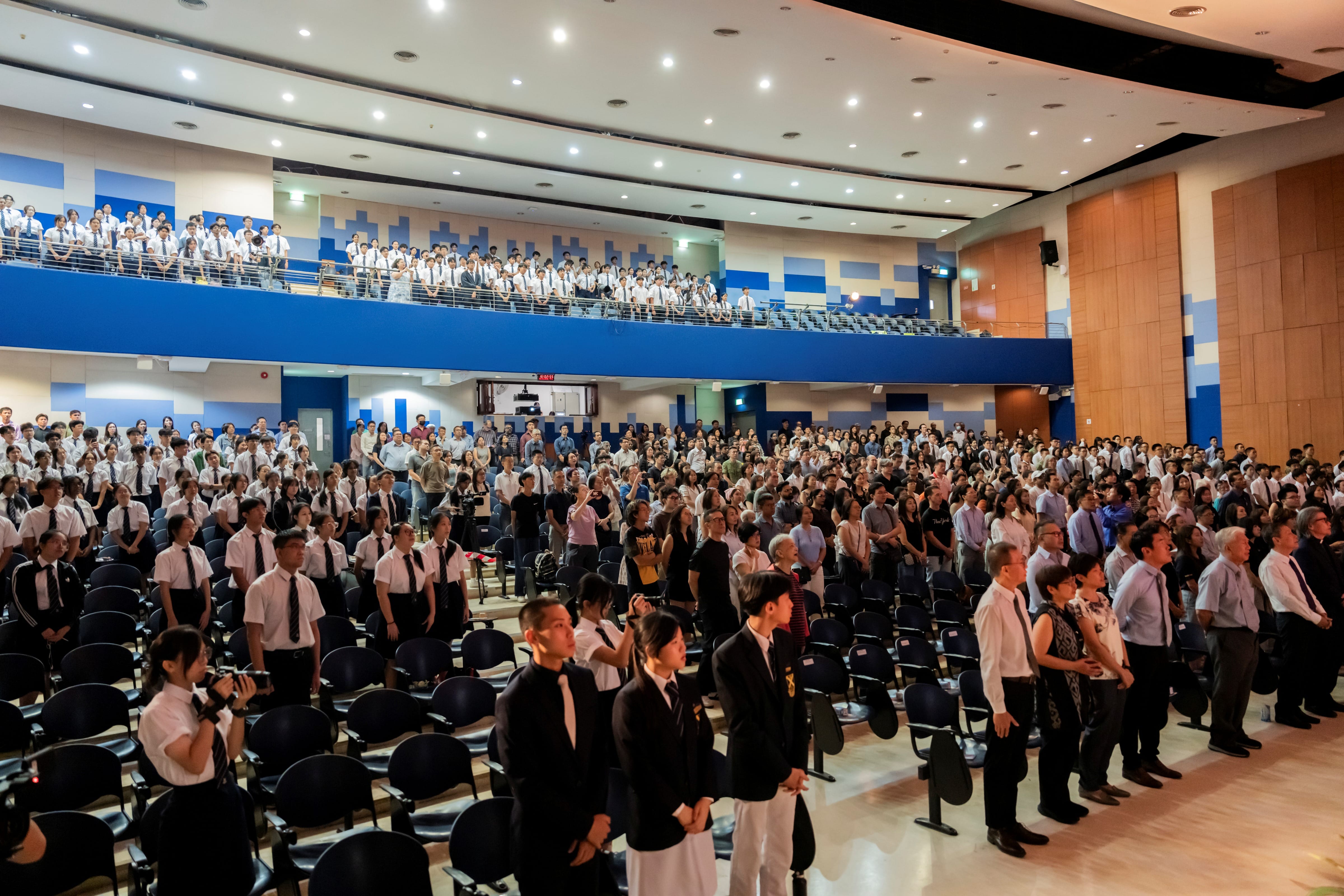 A packed audience of students, staff, and guests standing in a large multi-tiered auditorium during a school event.